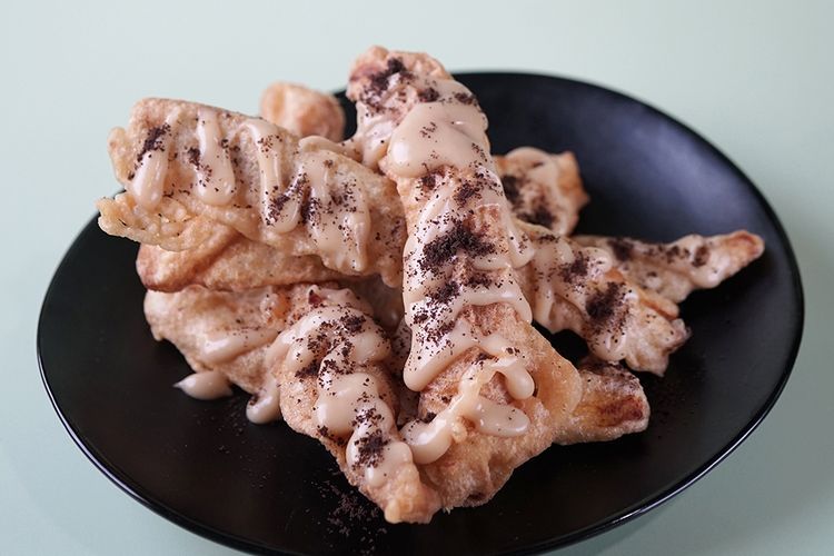 Sebuah porsi tiramisu pisang disajikan dalam piring putih, ditaburi dengan bubuk kakao dan dihiasi dengan irisan pisang di atasnya